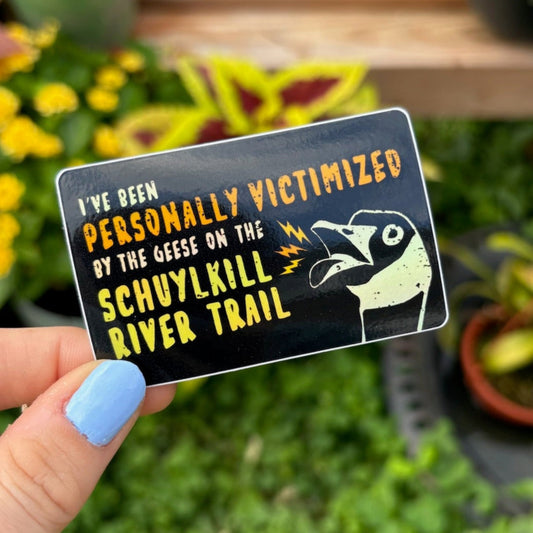 Hand holding a sticker with text saying "I've been personally victimized by the geese on the Schuylkill River Trail" with a goose hissing