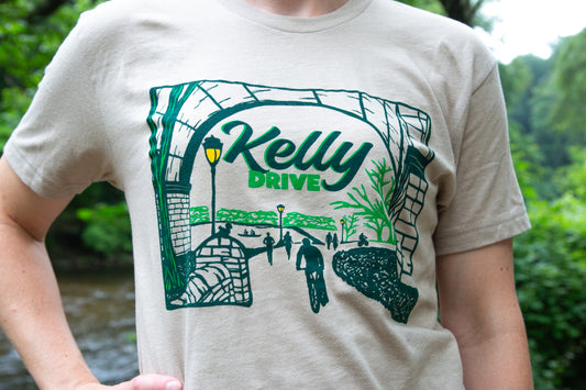 Person wearing a beige t-shirt with 'Kelly Drive' graphic design outdoors.