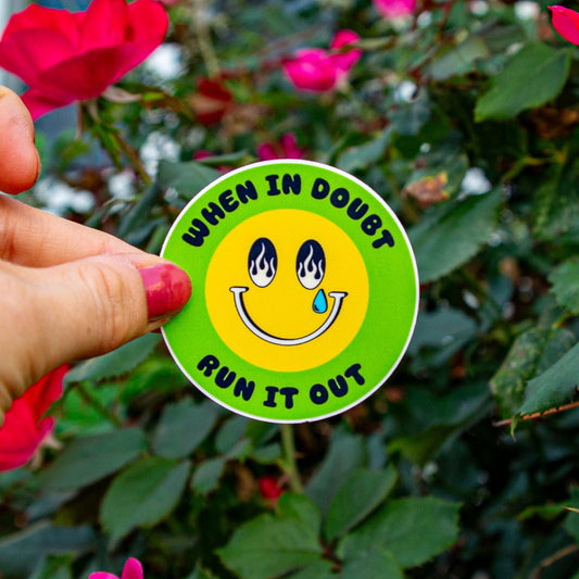 Hand holding a sticker with a smiley face and text 'When in Doubt Run it Out' in front of pink flowers and green leaves