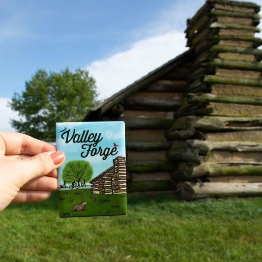 Hand holding a 'Valley Forge' magnet in front of a log cabin.