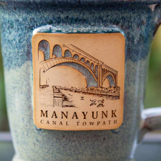 Close up on blue ceramic mug with 'Manayunk Canal Towpath' text and bridge illustration.