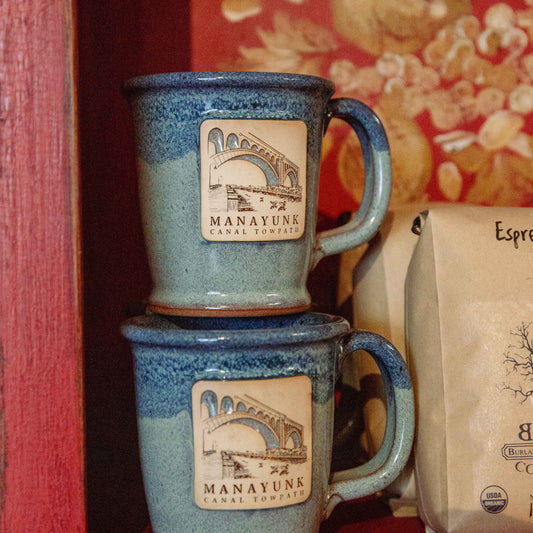 Two blue ceramic mugs with 'Manayunk Canal Towpath' clay illustration.