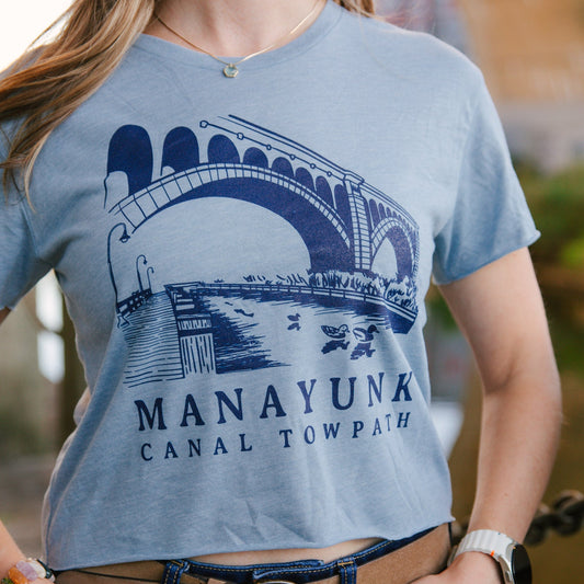 Person wearing a light blue t-shirt with a bridge graphic and 'Manayunk Canal Tow Path' text.