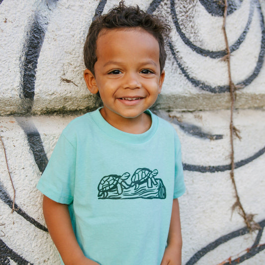Child wearing a light blue t-shirt with a turtle design, standing against a textured wall.