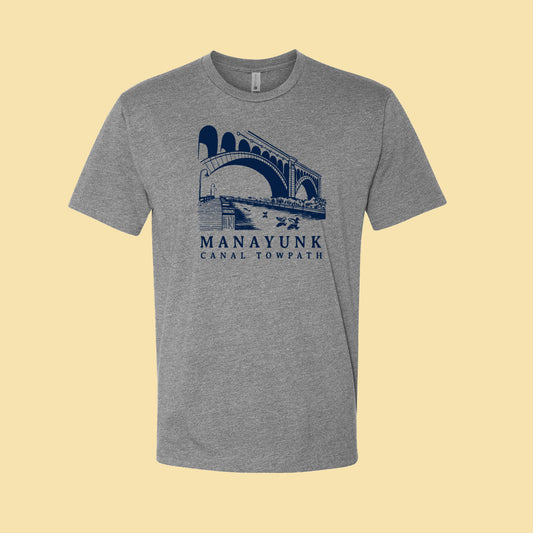 Heather gray t-shirt with a navy blue graphic of a trail and bridge and text 'Manayunk Canal Towpath' on a tan background.