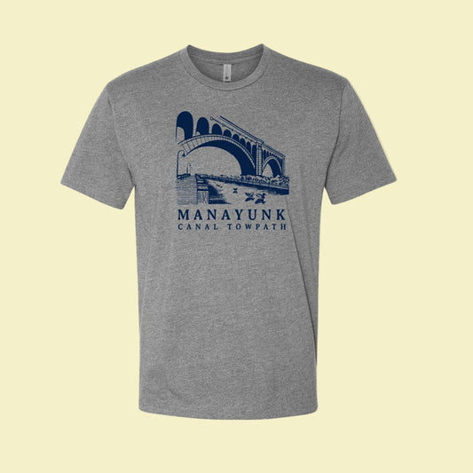 Heather gray t-shirt with a navy blue graphic of a trail and bridge and text 'Manayunk Canal Towpath' on a tan background.