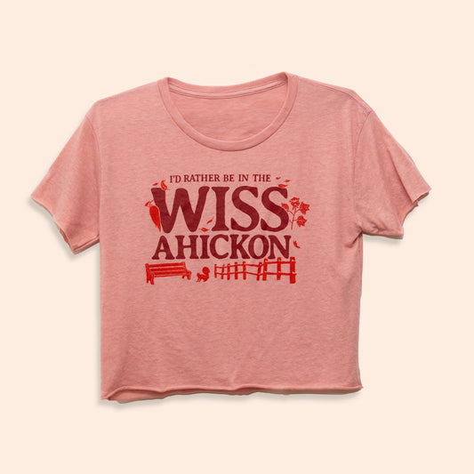 Pink crop top with red and maroon text 'I'd Rather Be in the Wissahickon' surrounded by a woodpecker, flowers, a bench, and fence on a beige background