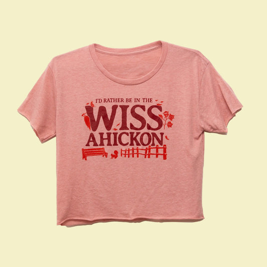 Pink crop top with red and maroon text 'I'd Rather Be in the Wissahickon' surrounded by a woodpecker, flowers, a bench, and fence on a beige background