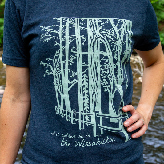 Young woman wearing a dark blue t-shirt with a light green fence and forest with text 'I'd rther be in the Wissahickon' while standing outdoors.