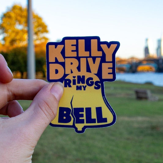 Hand holding a Librity bell shaped orange and blue sticker with 'Kelly Drive Rings against a blurred Kelly Drive background.
