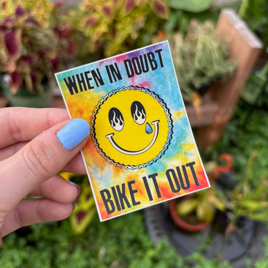 Hand holding a colorful sticker with a smiley face and text 'When in doubt, bike it out' against a garden background.