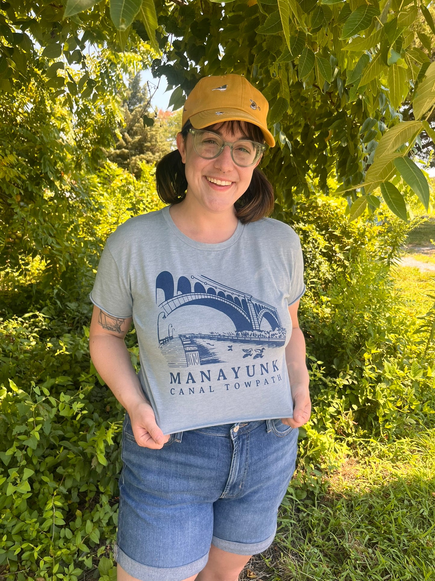 Person wearing a gray t-shirt with a bridge graphic and text, standing outdoors among greenery.