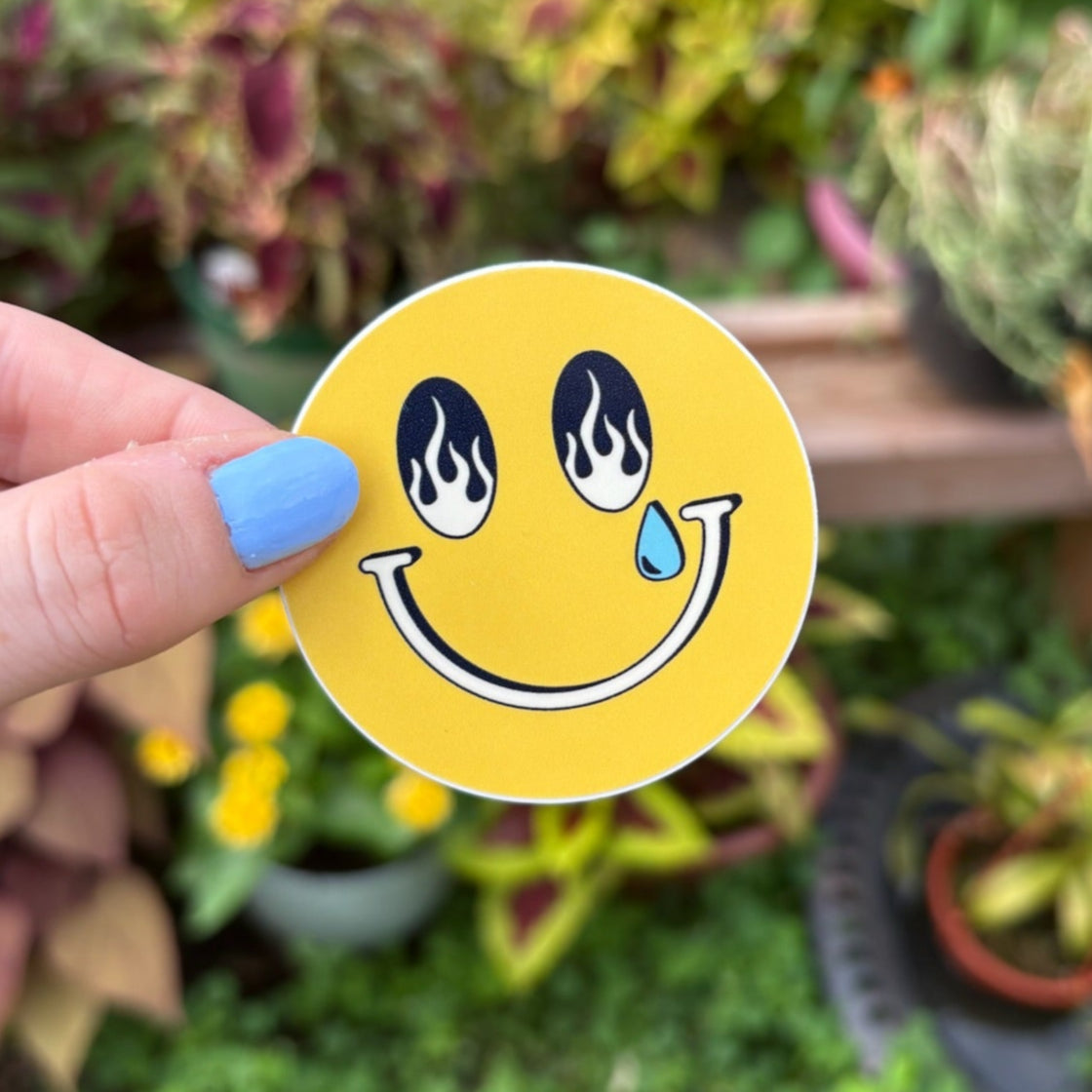 Hand holding a yellow smiley face sticker with a garden in the background. The smiley had fire design in it's eyes with one tear drop coming down.