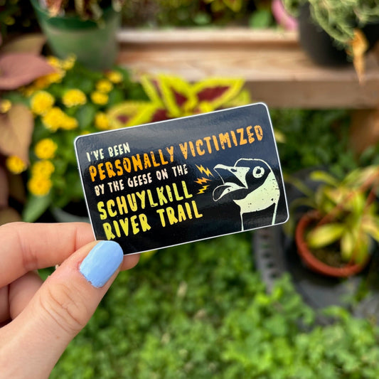 Hand holding a sticker with text saying "I've been personally victimized by the geese on the Schuylkill River Trail" with a goose hissing
