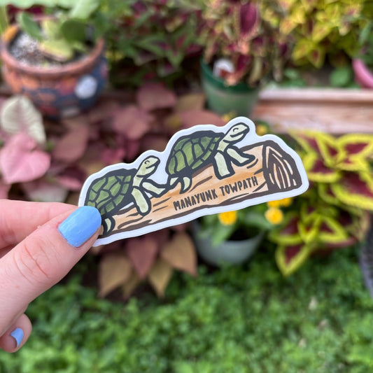Hand holding a sticker with two turtle sitting on a log with the text Manayunk Towpath on the log.