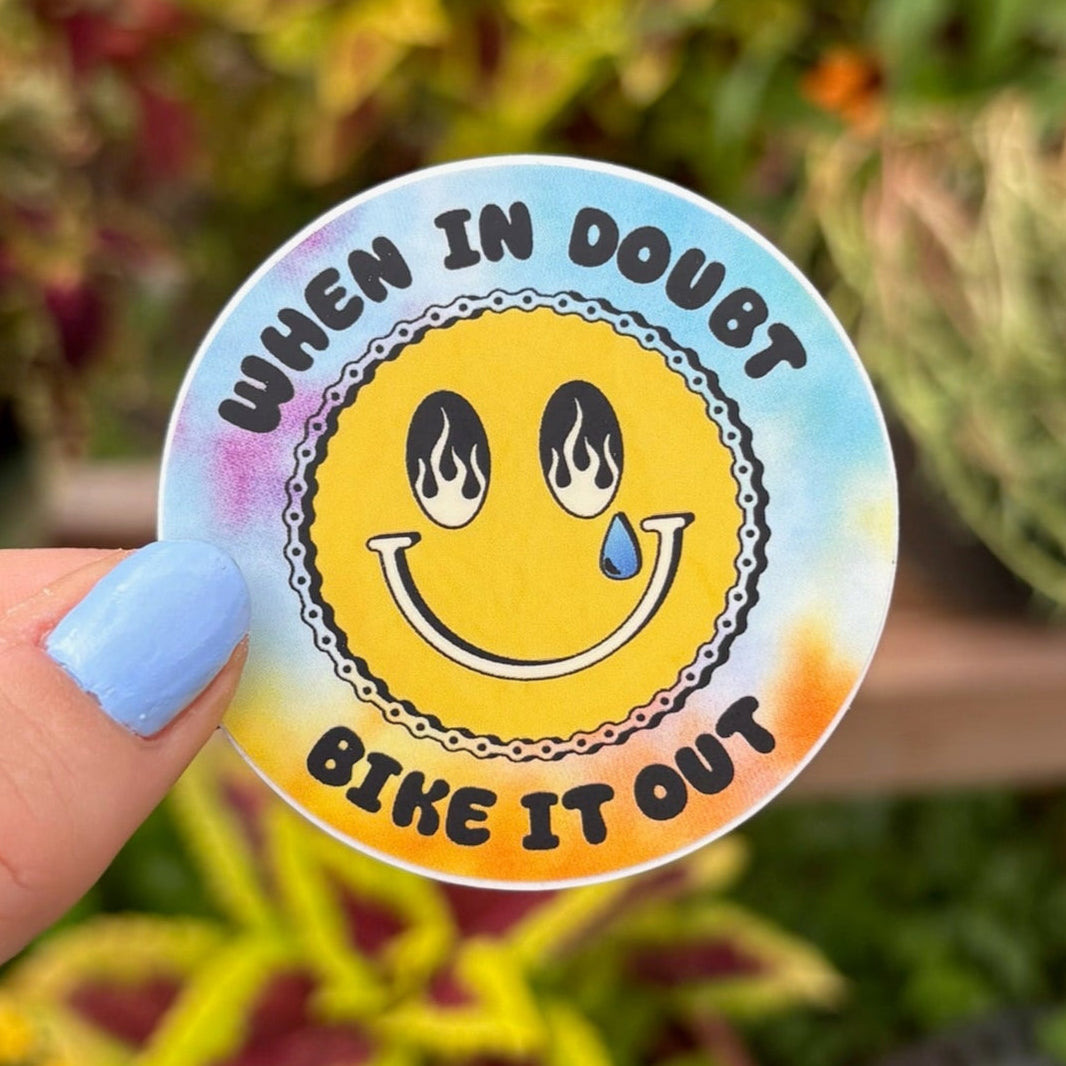 Sticker with smiley face and text 'When in doubt, bike it out' held in front of plants