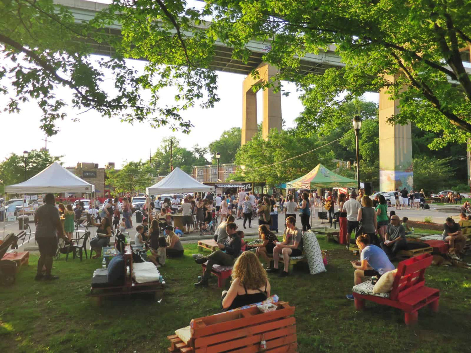 Outdoor event with people sitting on benches and chairs under trees, with a large architectural structure in the background.