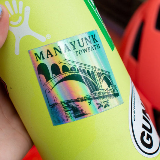A sticker with an illustration of a bridge over water, with the text 'MANAYUNK TOWPATH' across the top. The colors are vibrant, with a blue sky and green tones.