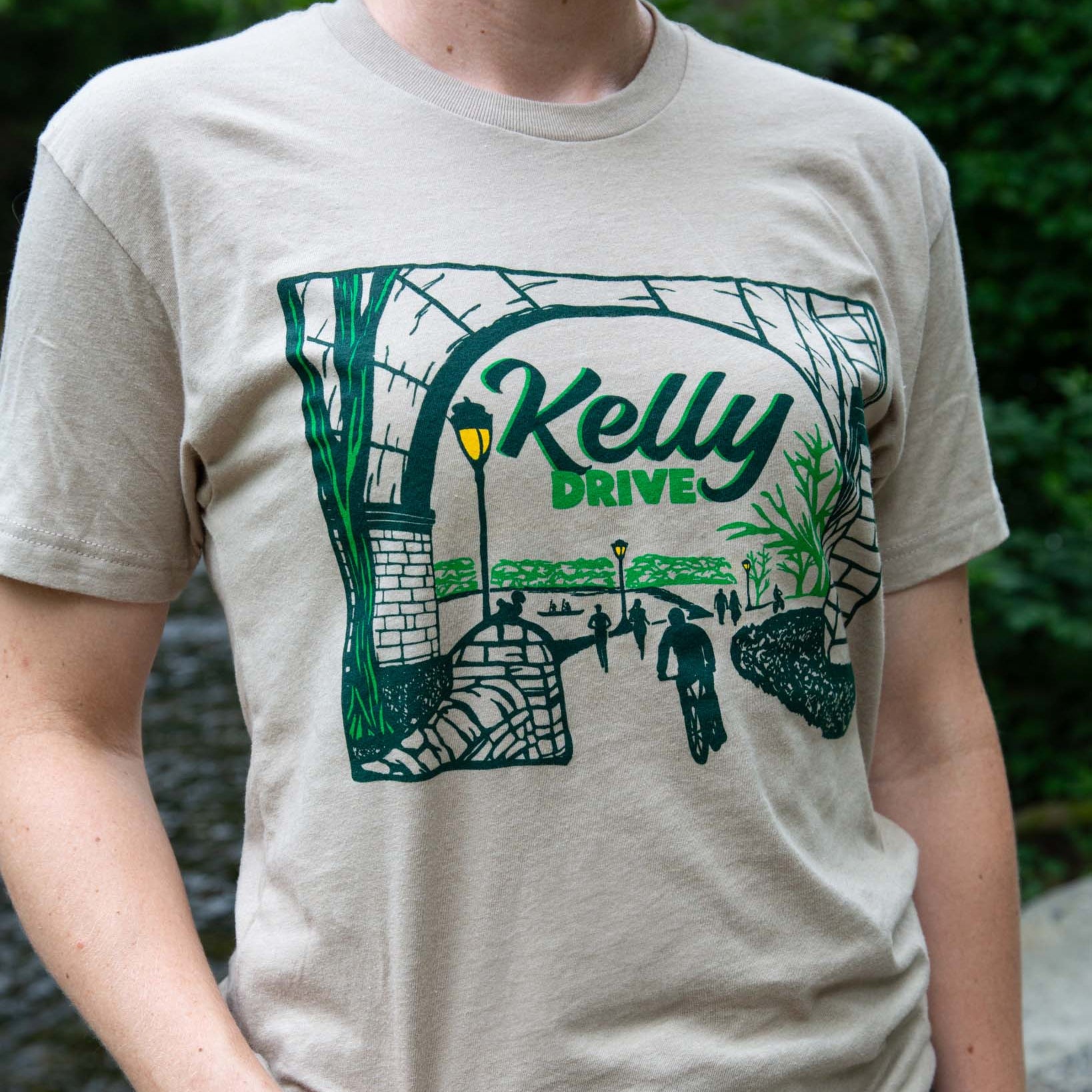 Person wearing a beige t-shirt with a green graphic design and 'Kelly Drive' text.