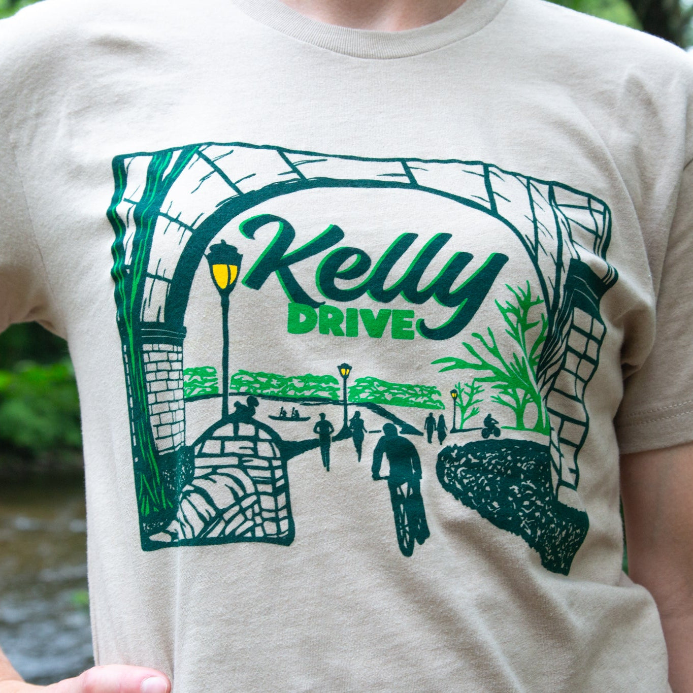 Beige t-shirt with 'Kelly Drive' design featuring a scenic view or bikers, runners and rowers on a trail.
