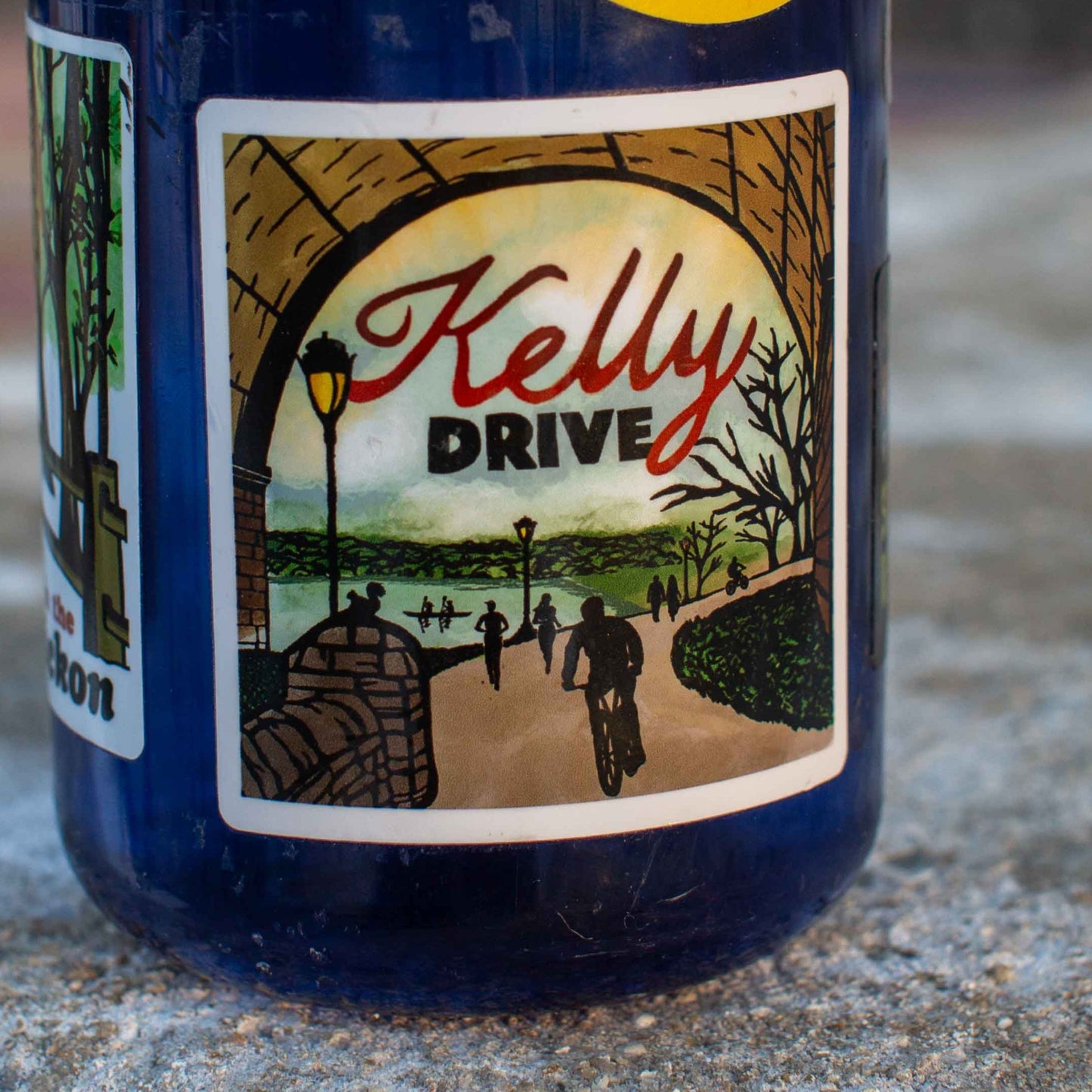 Bottle with a 'Kelly Drive' illustration sticker on a concrete surface. 