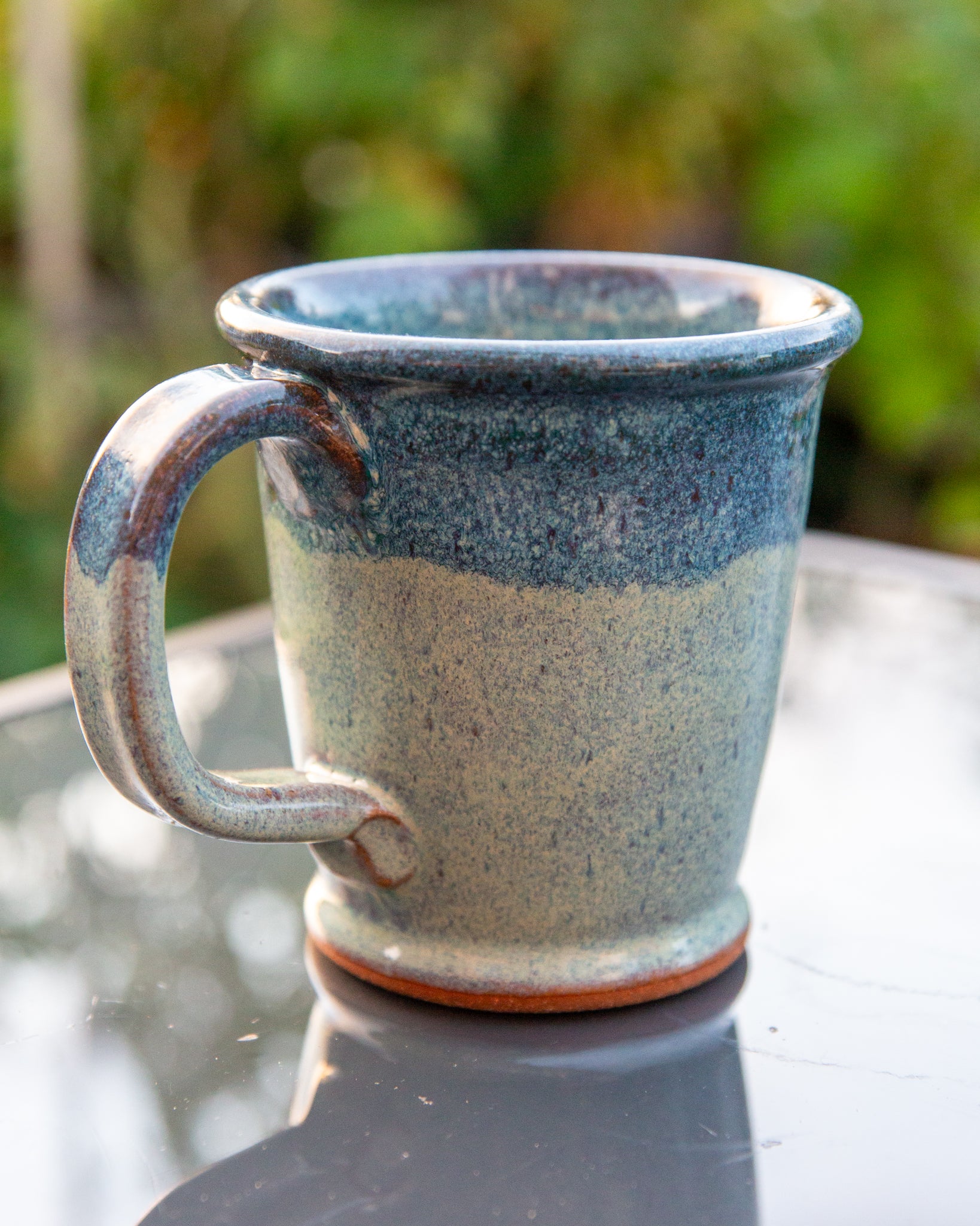 Handmade ceramic mug with blue and beige glaze on a reflective surface with blurred green background