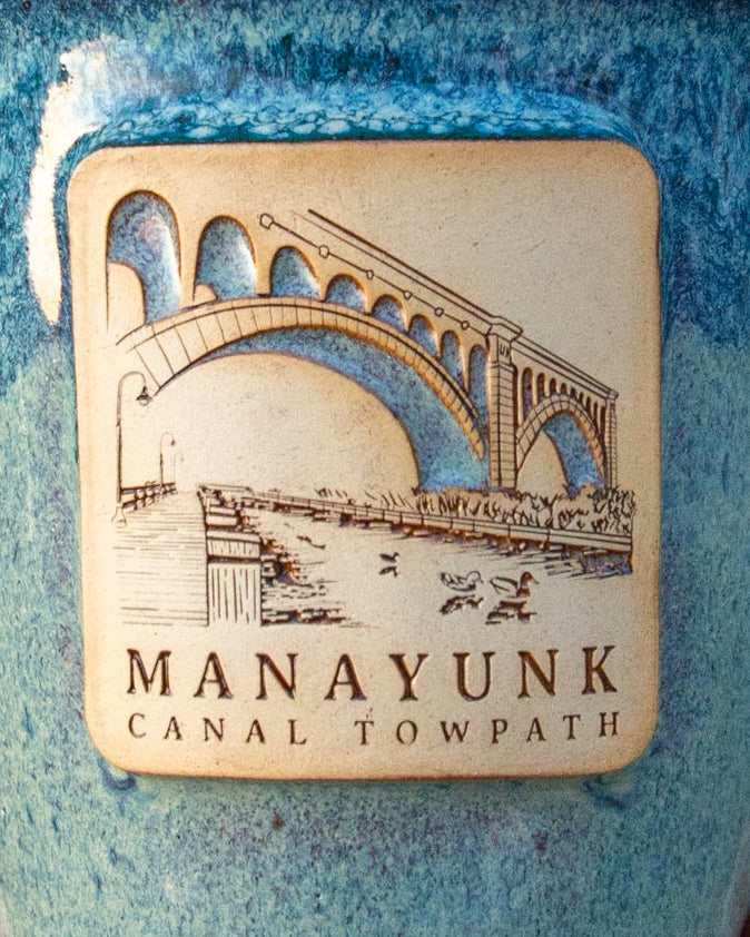 Blue ceramic mug with 'Manayunk Canal Towpath' design held in a hand against a green leafy background