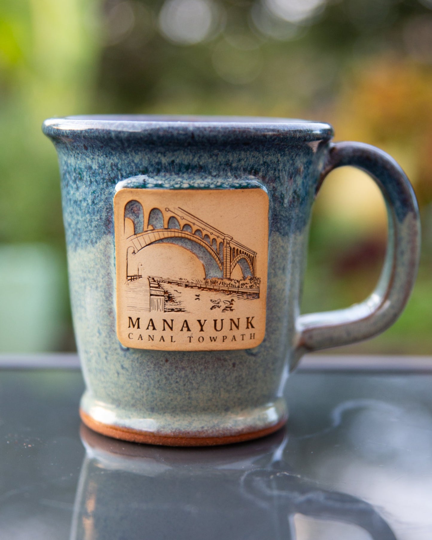 Blue ceramic mug featuring 'Manayunk Canal Towpath' on a reflective surface.