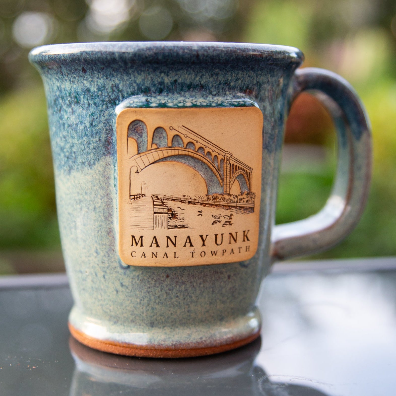 Blue ceramic mug with a bridge logo and 'Manayunk Canal Towpath' text on a reflective surface with blurred green background