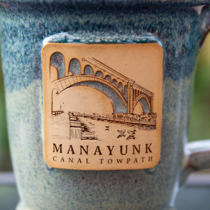 Close up on blue ceramic mug with 'Manayunk Canal Towpath' text and bridge illustration.