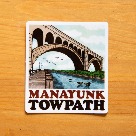 A sticker featuring an illustration of the Manayunk Towpath with a bridge, water, and birds in a stylized representation of the area on a wooden background.