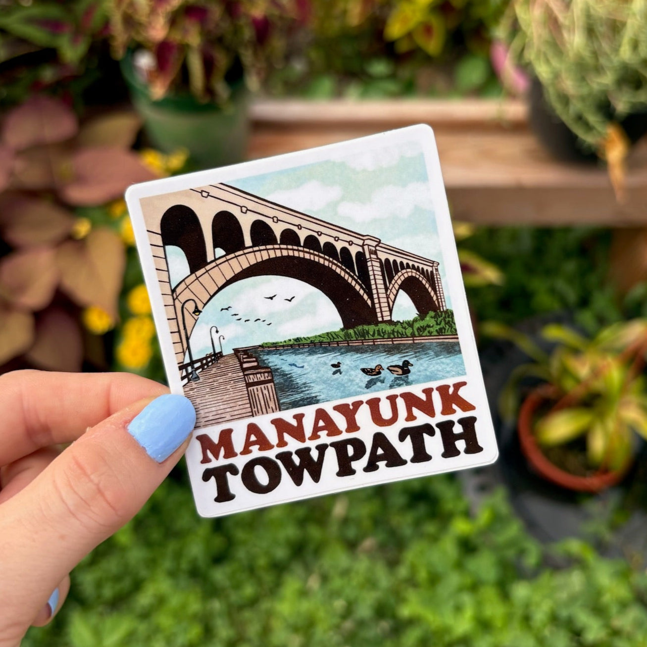 A sticker featuring an illustration of the Manayunk Towpath with a bridge, water, and birds in a stylized representation of the area. 