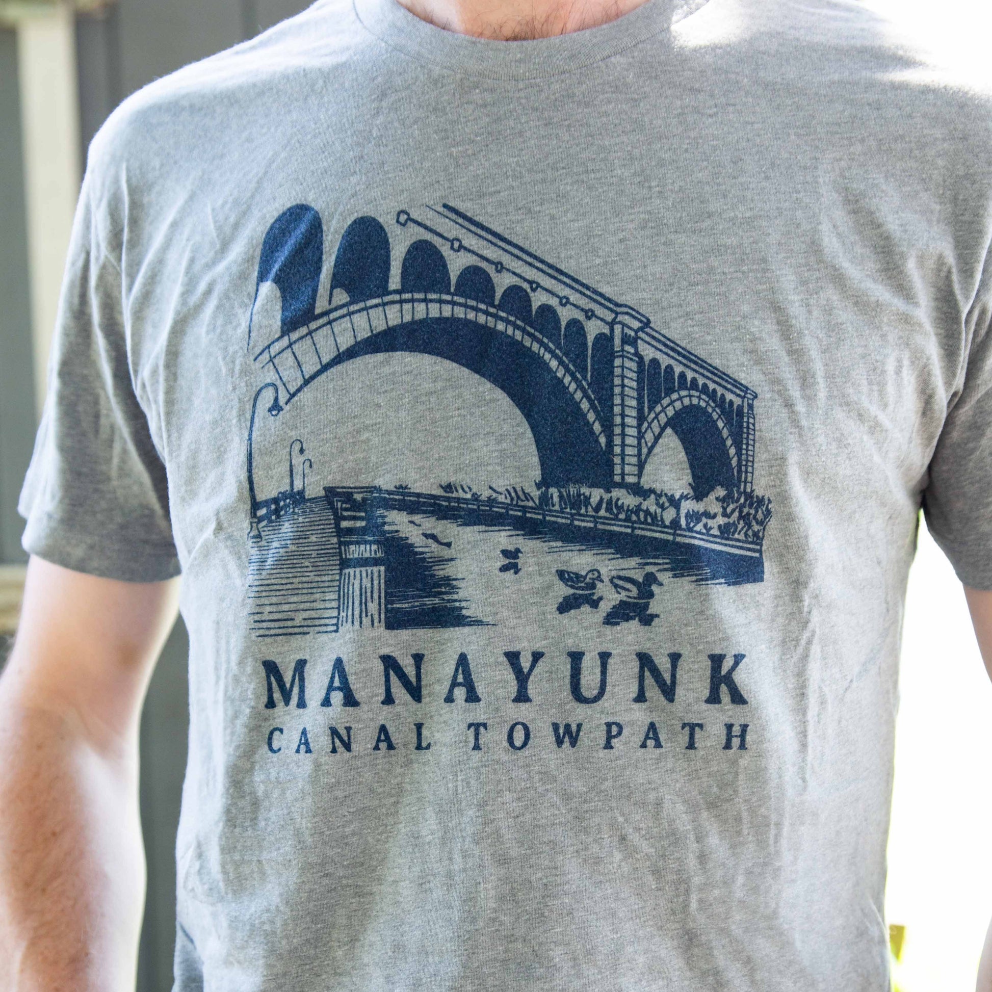 A person wearing a gray t-shirt with 'MANAYUNK CANAL TOWPATH' printed in blue text, featuring an illustration of a bridge and canal.