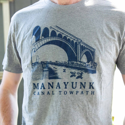 A person wearing a gray t-shirt with 'MANAYUNK CANAL TOWPATH' printed in blue text, featuring an illustration of a bridge and canal.