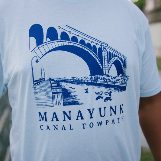 Light blue t-shirt with a blue graphic of a trail and bridge and text 'Manayunk Canal Towpath' on a person outdoors.