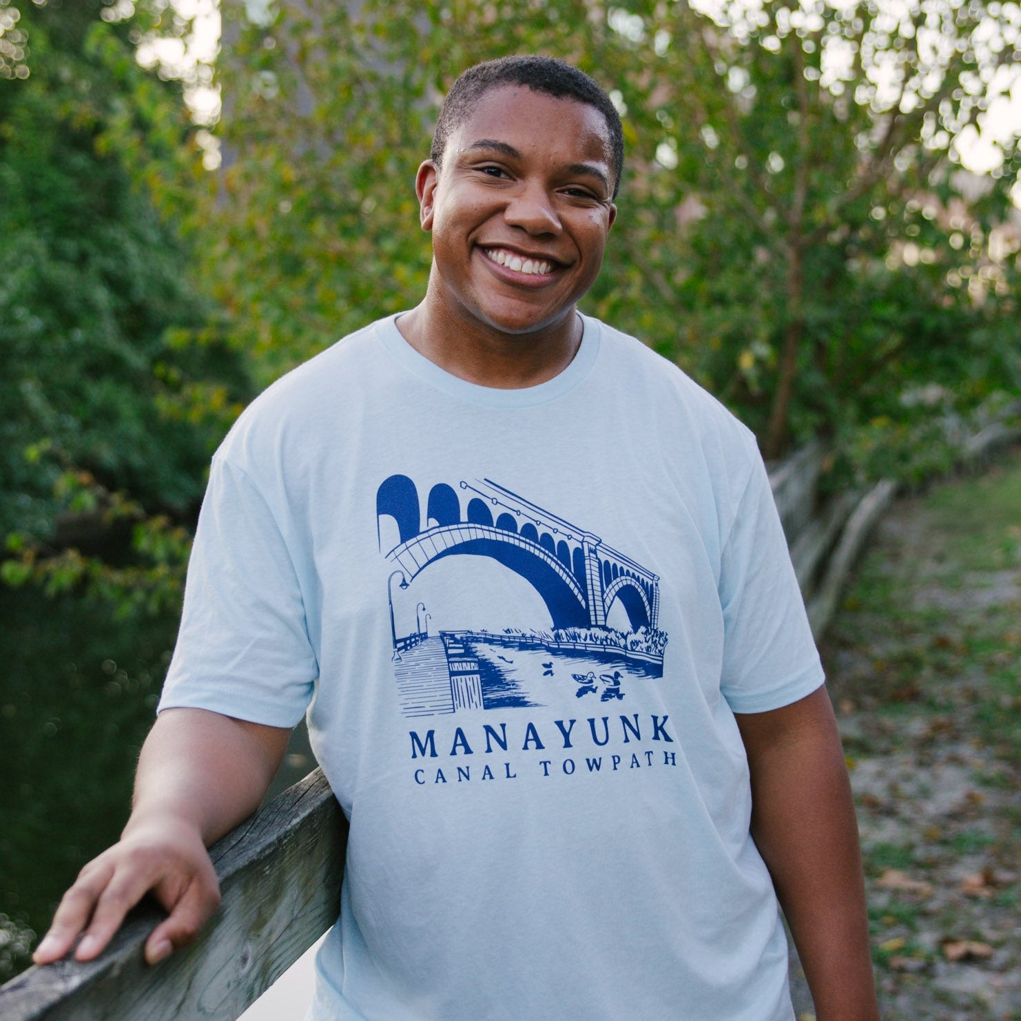 Person wearing a light blue t-shirt with 'Manayunk Canal Towpath' design, standing outdoors.