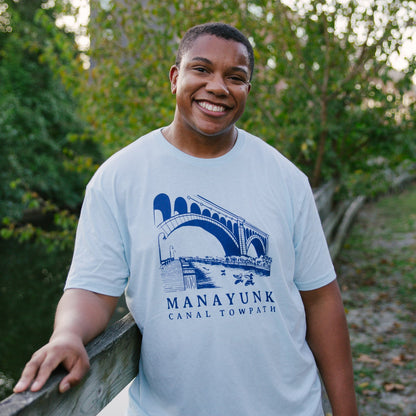 Person wearing a light blue t-shirt with 'Manayunk Canal Towpath' design, standing outdoors.