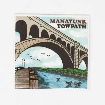 A fridge magnet featuring an illustration of the Manayunk Towpath with a bridge, water, and birds in a stylized representation of the area.
