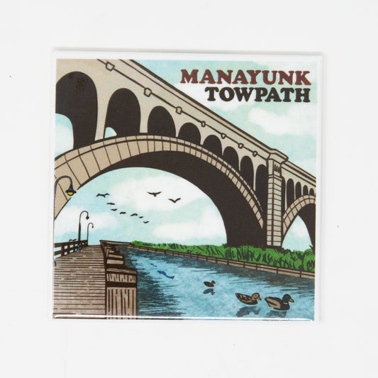 A fridge magnet featuring an illustration of the Manayunk Towpath with a bridge, water, and birds in a stylized representation of the area.