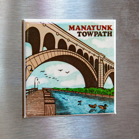 A fridge magnet featuring an illustration of the Manayunk Towpath with a bridge, water, and birds in a stylized representation of the area on a metalic surface.