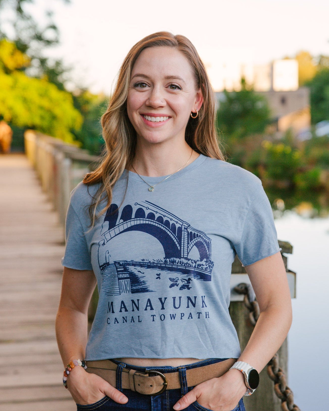 Woman wearing a blue t-shirt with a bridge graphic and text, standing on a wooden bridge over a canal.