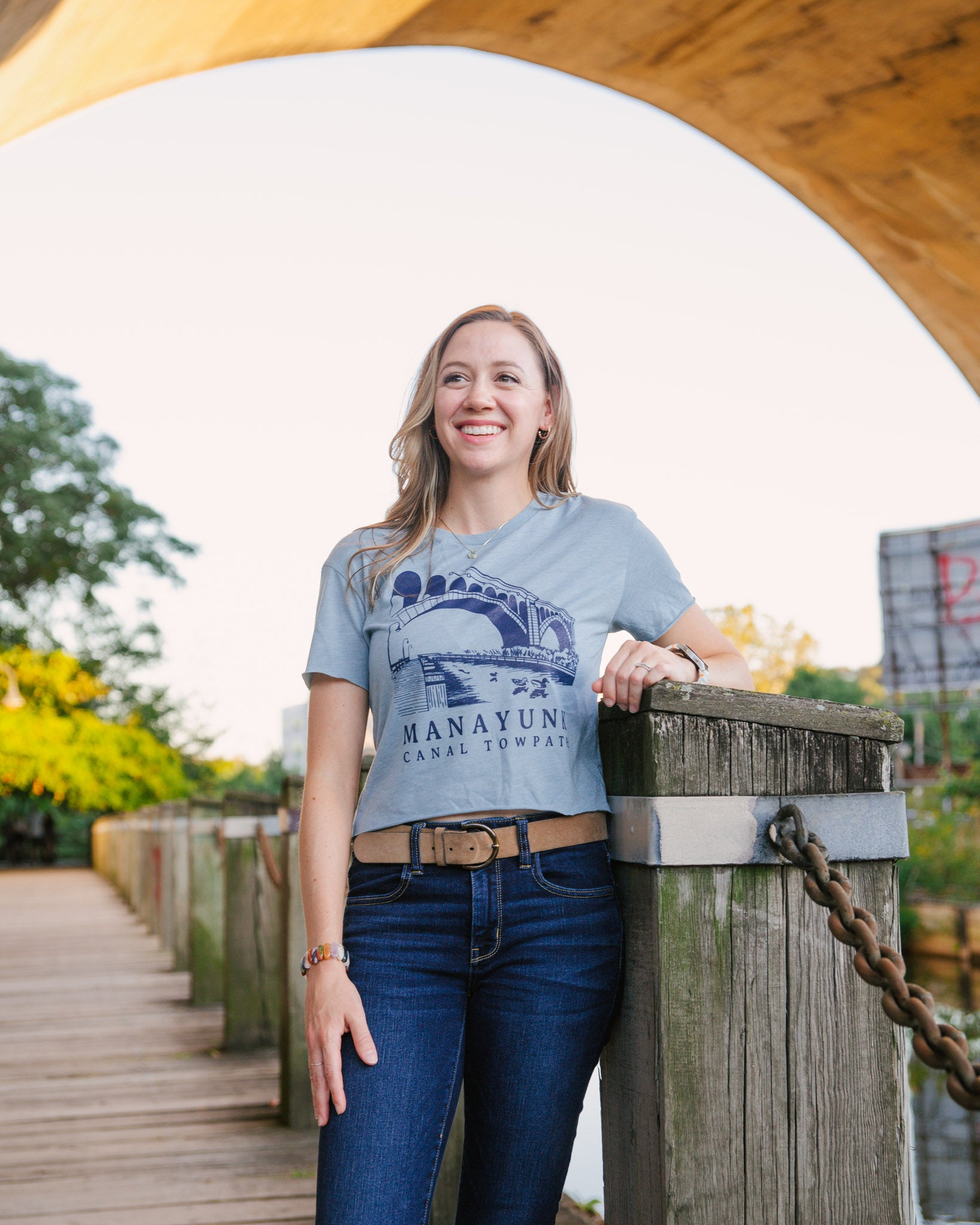 Woman wearing a blue t-shirt with 'Manayunk Canal Towpath' design, standing by a wooden post.