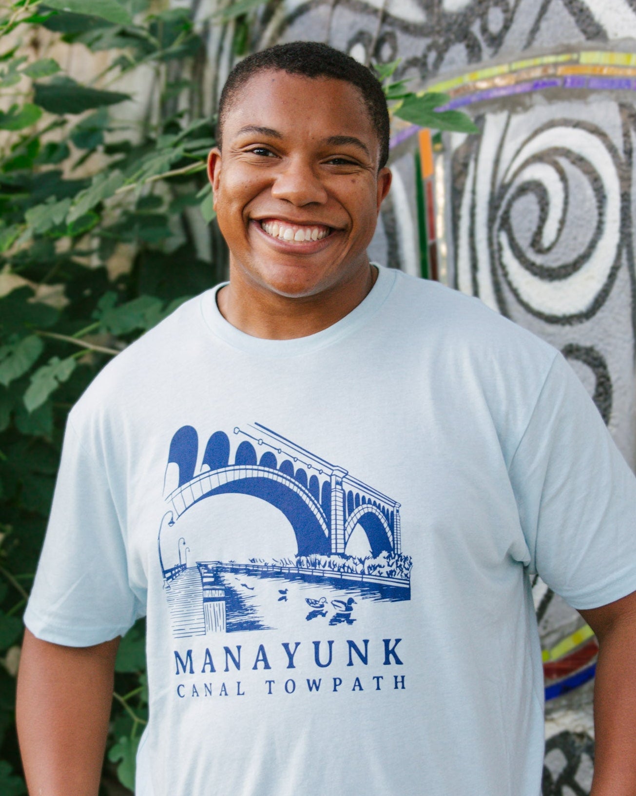 Person wearing a light blue t-shirt with 'Manayunk Canal Towpath' design, standing in front of a decorative wall.