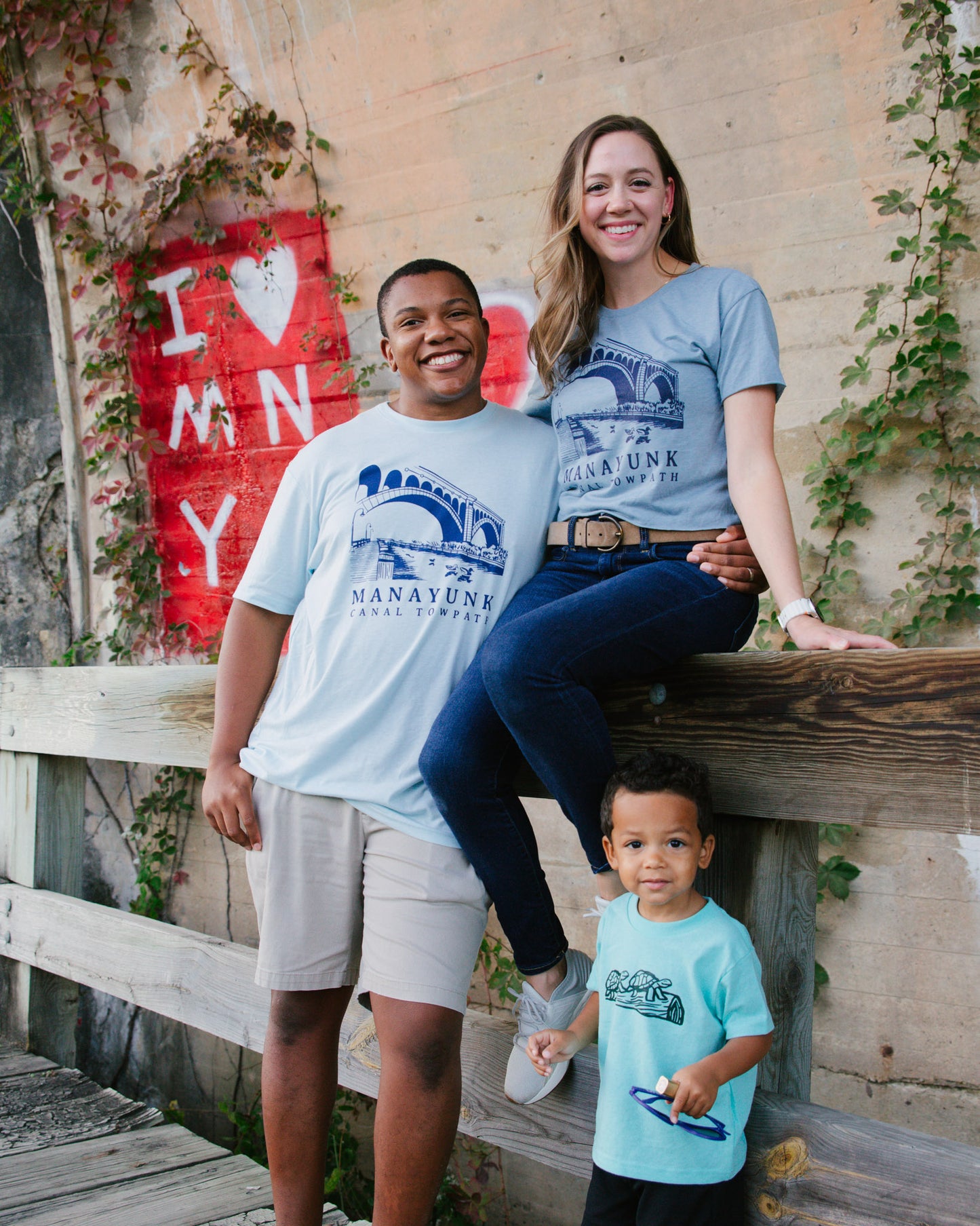 Two adults and a child wearing Manayunk t-shirts standing on a wooden bridge with a red 'I ❤️ MN' sign in the background.