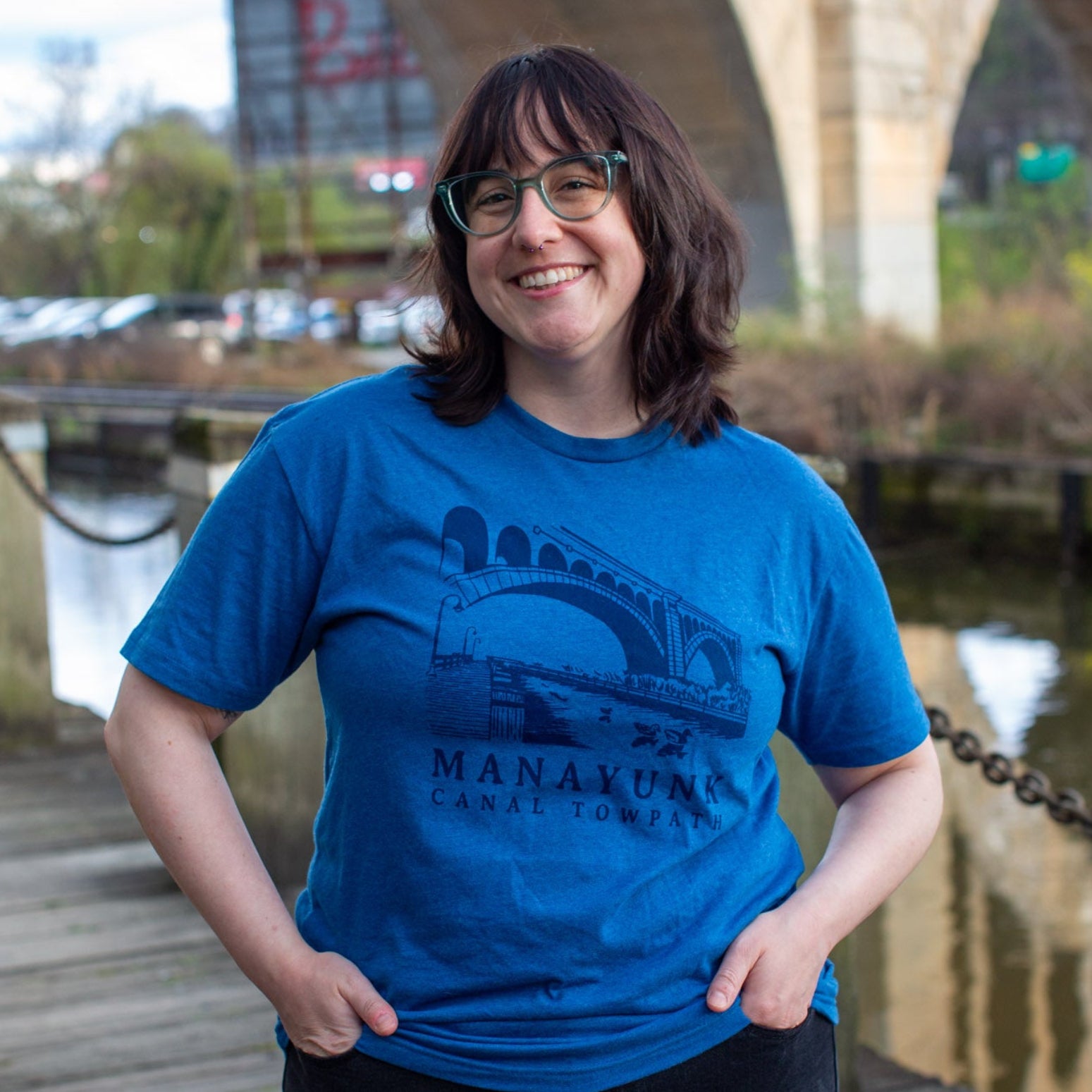 Person wearing a dark blue t-shirt with 'Manayunk Canal Towpath' design, standing in front of a bridge outside. 