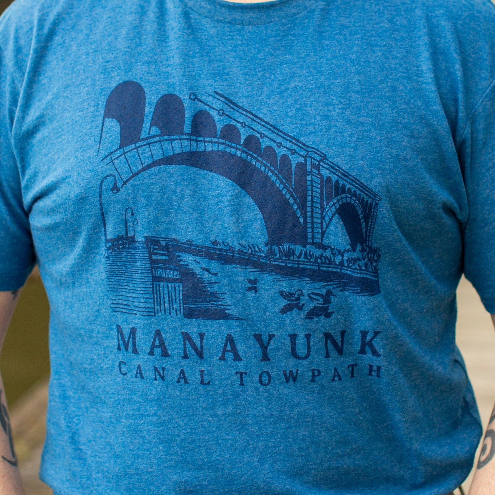 A person wearing a gray t-shirt with 'MANAYUNK CANAL TOWPATH' printed in blue text, featuring an illustration of a bridge and canal.