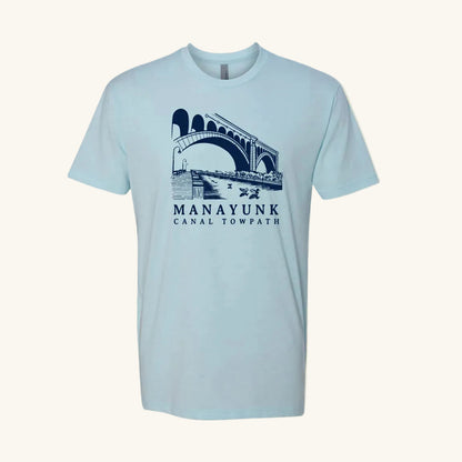Light blue t-shirt with a navy blue graphic of a trail and bridge and text 'Manayunk Canal Towpath' on a tan background.