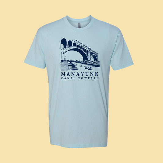 Light blue t-shirt with a navy blue graphic of a trail and bridge and text 'Manayunk Canal Towpath' on a tan background.