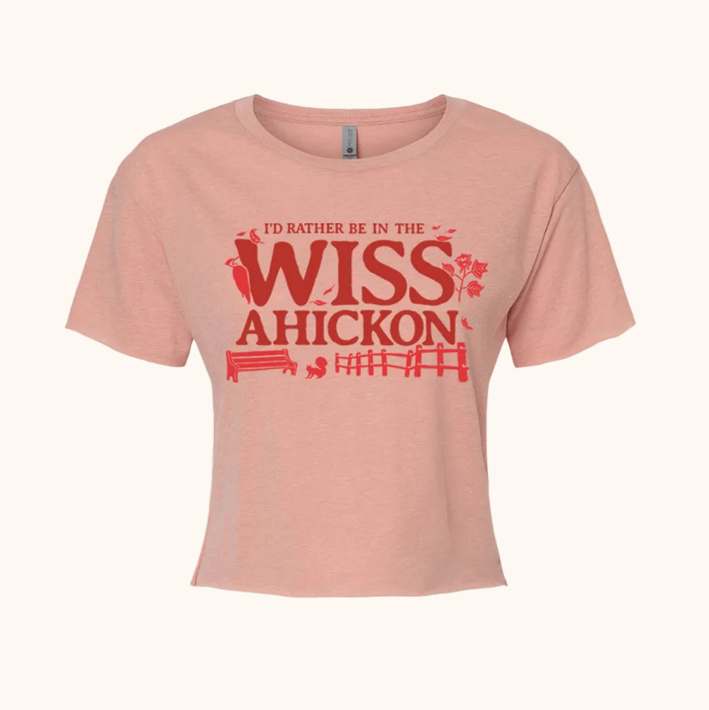 Pink crop top with red and maroon text 'I'd Rather Be in the Wissahickon' surrounded by a woodpecker, flowers, a bench, and fence on a beige background. 