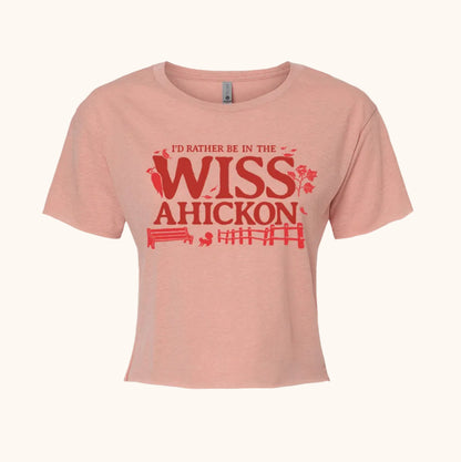 Pink crop top with red and maroon text 'I'd Rather Be in the Wissahickon' surrounded by a woodpecker, flowers, a bench, and fence on a beige background. 